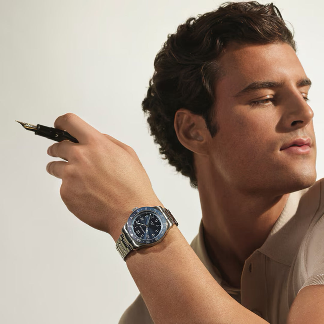 A man with dark hair in a beige shirt holds a fountain pen and looks to the side. On his left wrist, he wears the elegant Montblanc 1858 GMT 0 Oxygen Automatic 42mm Watch. The softly lit background remains neutral.