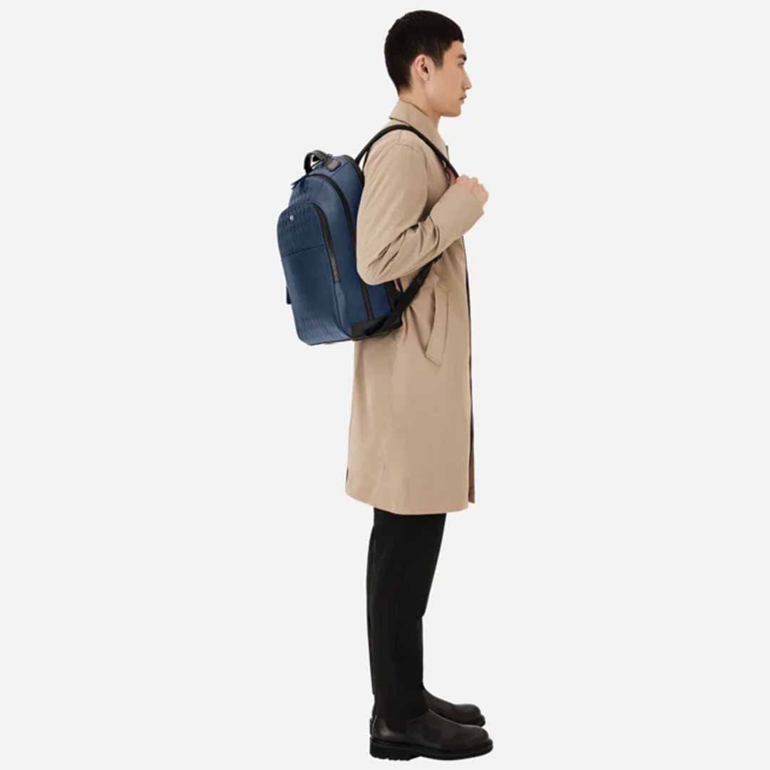 A person in a beige trench coat and black pants stands sideways, highlighting the Montblanc Extreme 3.0 Blue Leather Backpack with an embossed full-grain leather finish. The blue accessory features a padded laptop compartment, ideal for modern needs against the plain white backdrop.