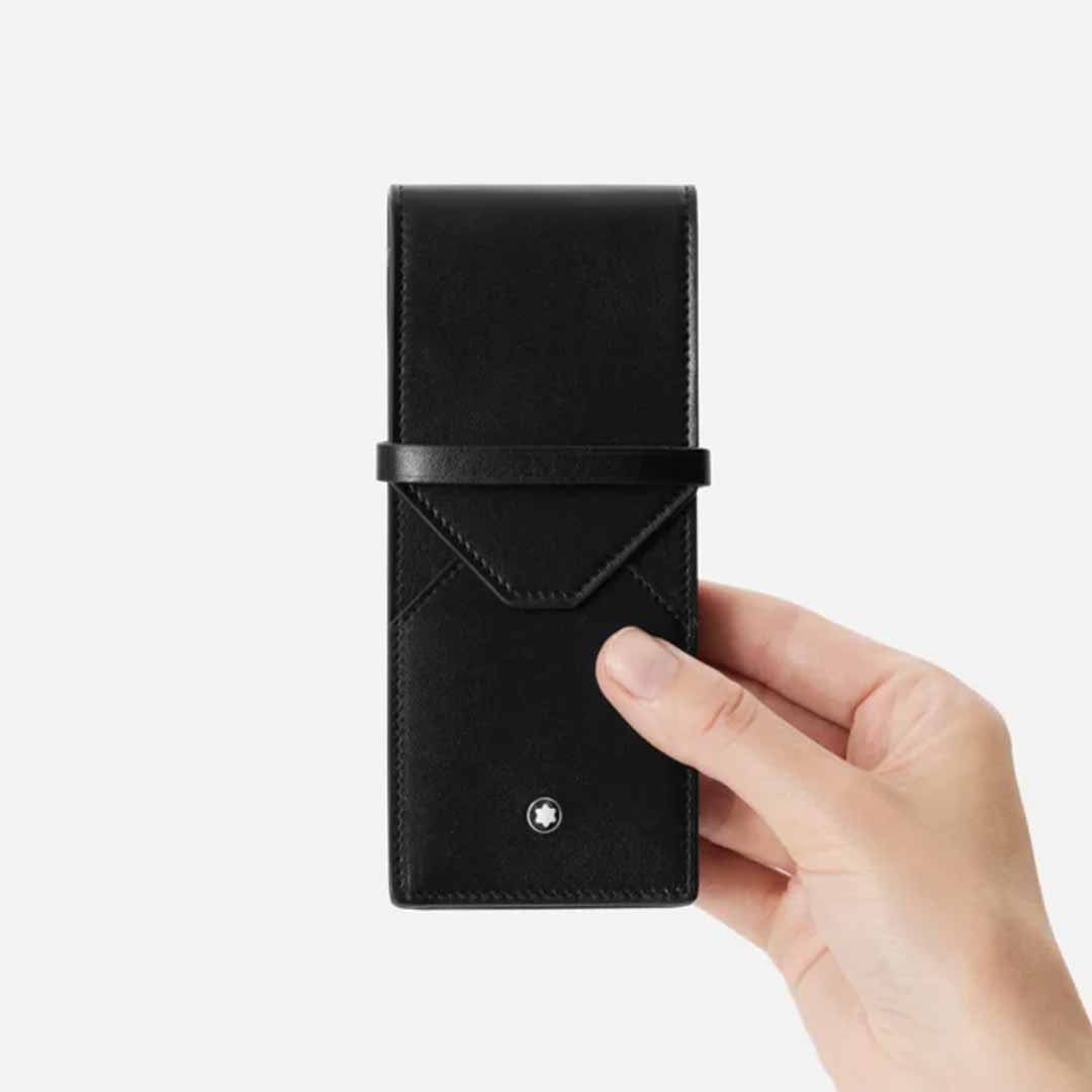 A hand holds the Montblanc Meisterstück 3-Pen Leather Pouch, showcasing a sleek black leather design with a minimalist aesthetic. The iconic Montblanc emblem is prominently displayed near the bottom. This luxury pen pouch features a smooth texture and an envelope-like flap closure, all set against a plain white background.