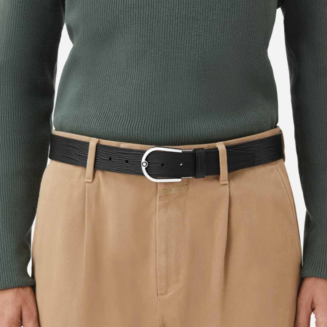 A person wears a dark green ribbed sweater tucked into beige pleated pants, cinched with the Montblanc Horseshoe 35mm Reversible Leather Belt featuring a silver palladium-finish buckle. The minimalistic style is highlighted against a plain white background.