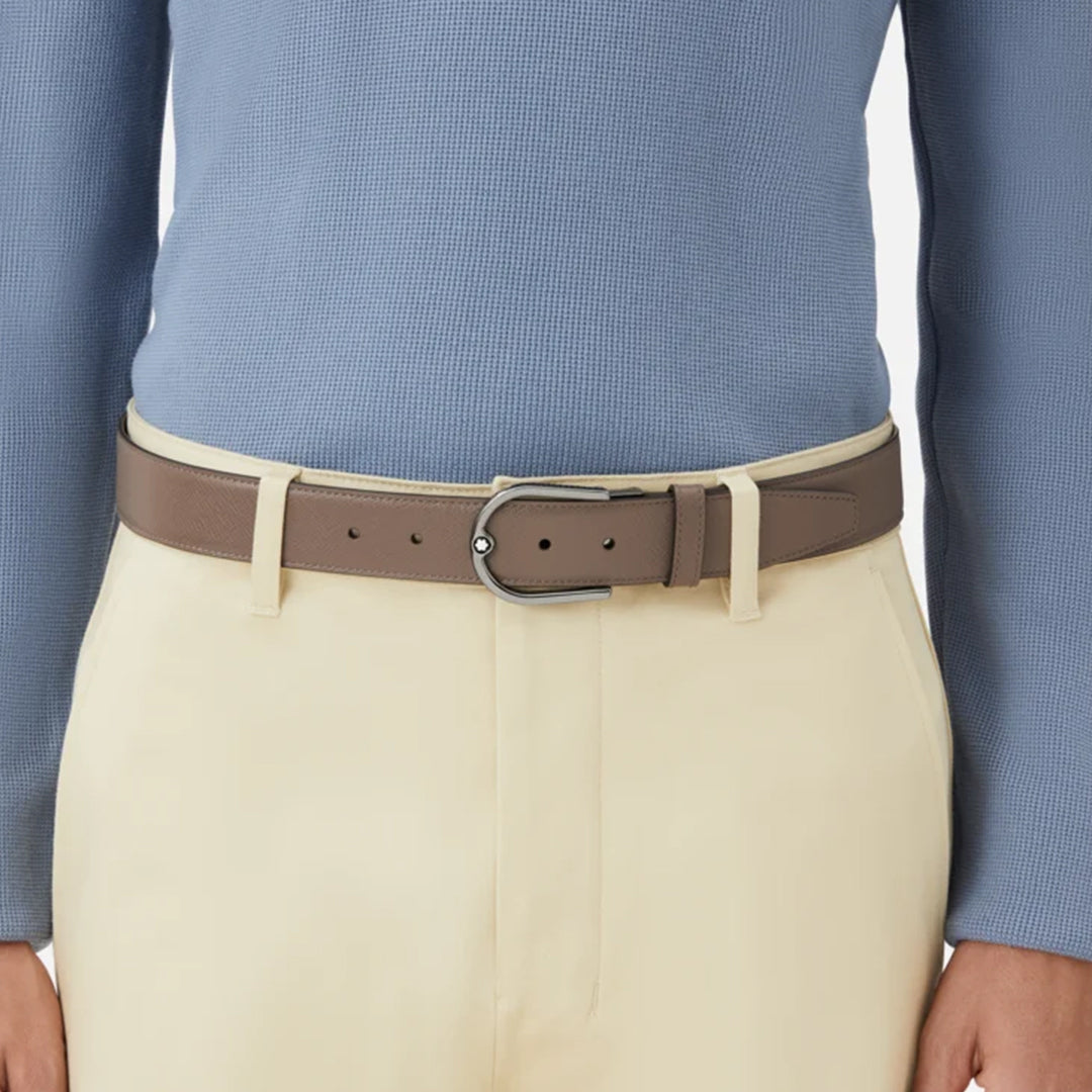 A person wears a light blue long-sleeve shirt and cream-colored pants, complemented by a neatly tucked Montblanc Horseshoe Buckle Saffiano Printed Mastic 35mm Reversible Leather Belt. The outfit achieves a clean, smart-casual look with the belt’s silver buckle adding sophistication.
