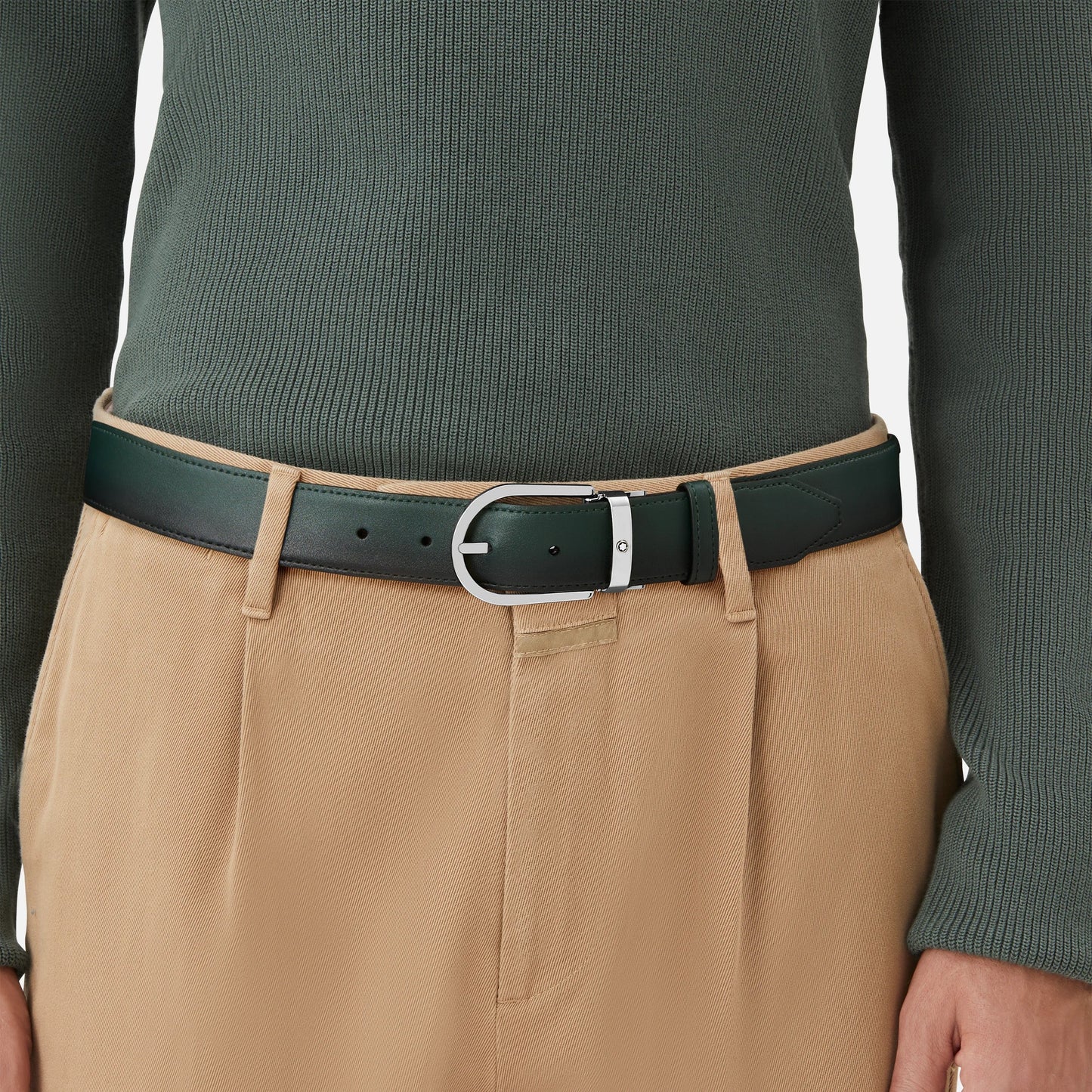 A person is wearing beige pants and a dark British green ribbed long-sleeve shirt, accessorized with the Montblanc Horseshoe Buckle Sfumato British Green 35mm Leather Belt. The image focuses on the midsection, highlighting the clothing and belt details.
