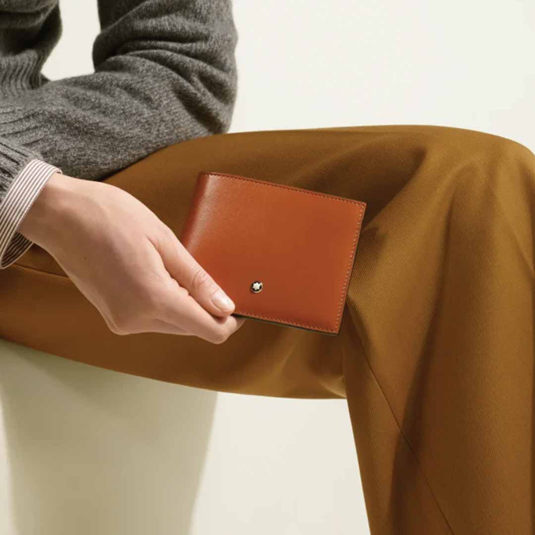 A person wearing a gray sweater and brown pants is holding an elegant, rust-colored Montblanc Meisterstück 6cc Leather Wallet, featuring a small metallic logo and designed to neatly fit six credit cards.