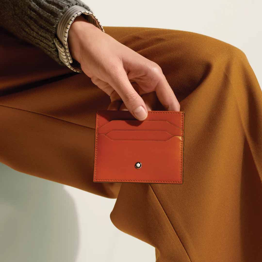 An individual holds a Montblanc Meisterstuck 6cc Leather Card Holder in orange leather against a backdrop of burnt orange fabric. The person is dressed in a gray sweater, with their wrist adorned by a watch. The card holder prominently features the Montblanc emblem and several meticulously crafted card slots.