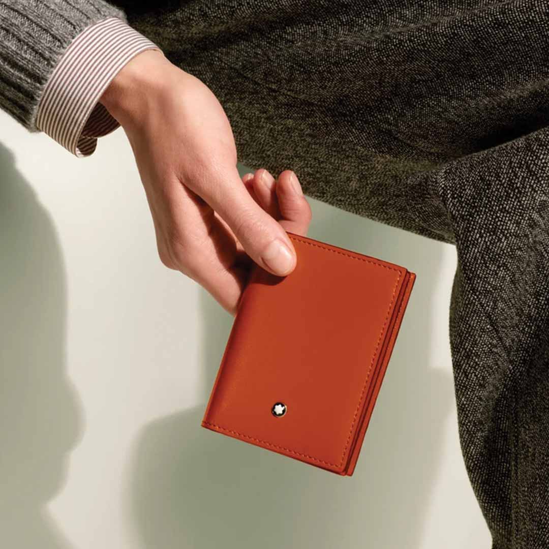 A person wearing a gray sweater and a pinstriped shirt holds an orange Montblanc Meisterstuck 4cc Leather Card Holder with a small white emblem, set against a greenish background.