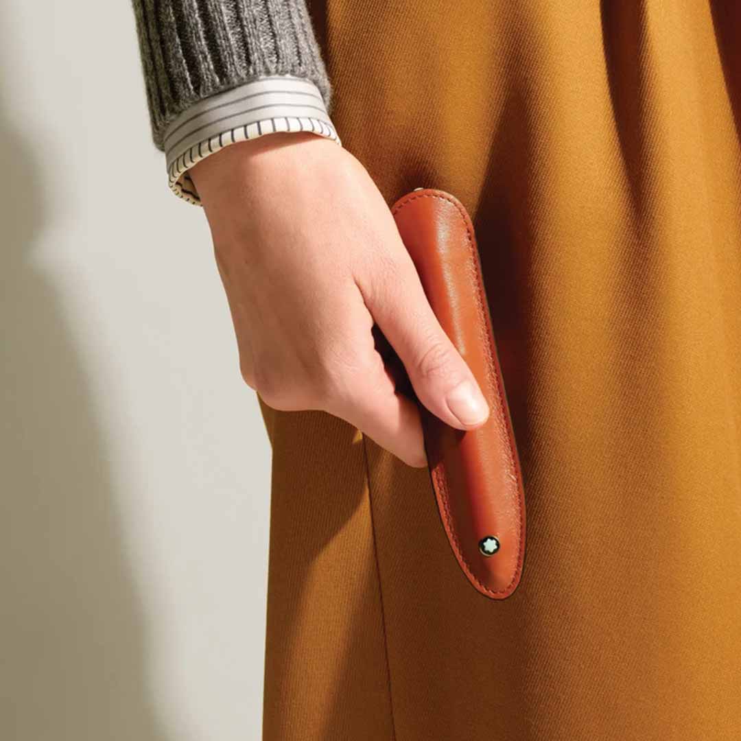 A person wearing a gray sweater and striped shirt holds the sophisticated Montblanc Meisterstück Brown 1 Pen Sleeve, famed for its slim profile and button closure. The sleeve is set against a mustard-colored fabric background.