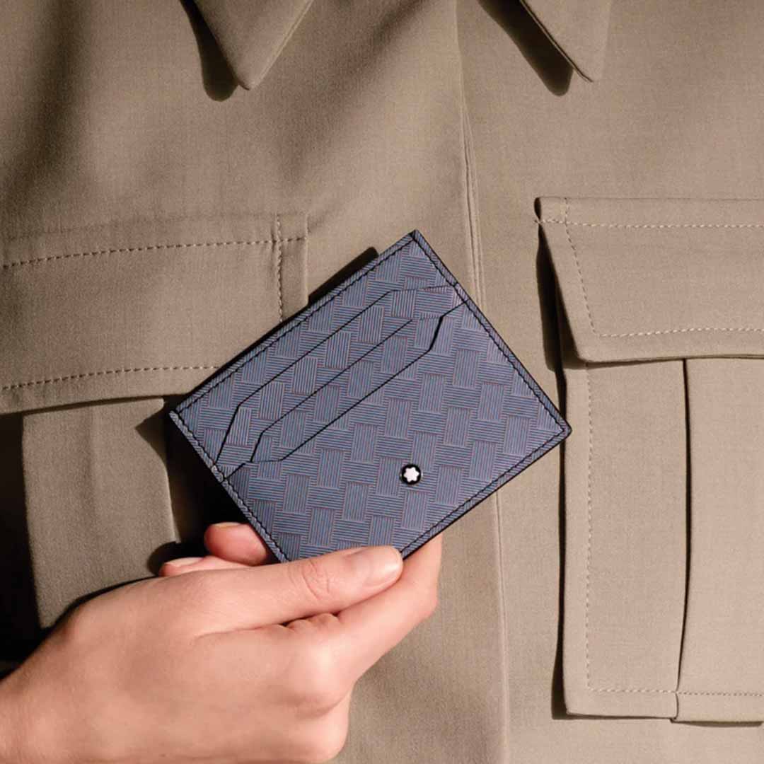 A person holds a Montblanc Extreme 3.0 6cc Leather Card Holder in a stylish blue, featuring a textured geometric pattern with a small logo at the center. They wear a beige jacket with large pockets, enhancing their chic ensemble.