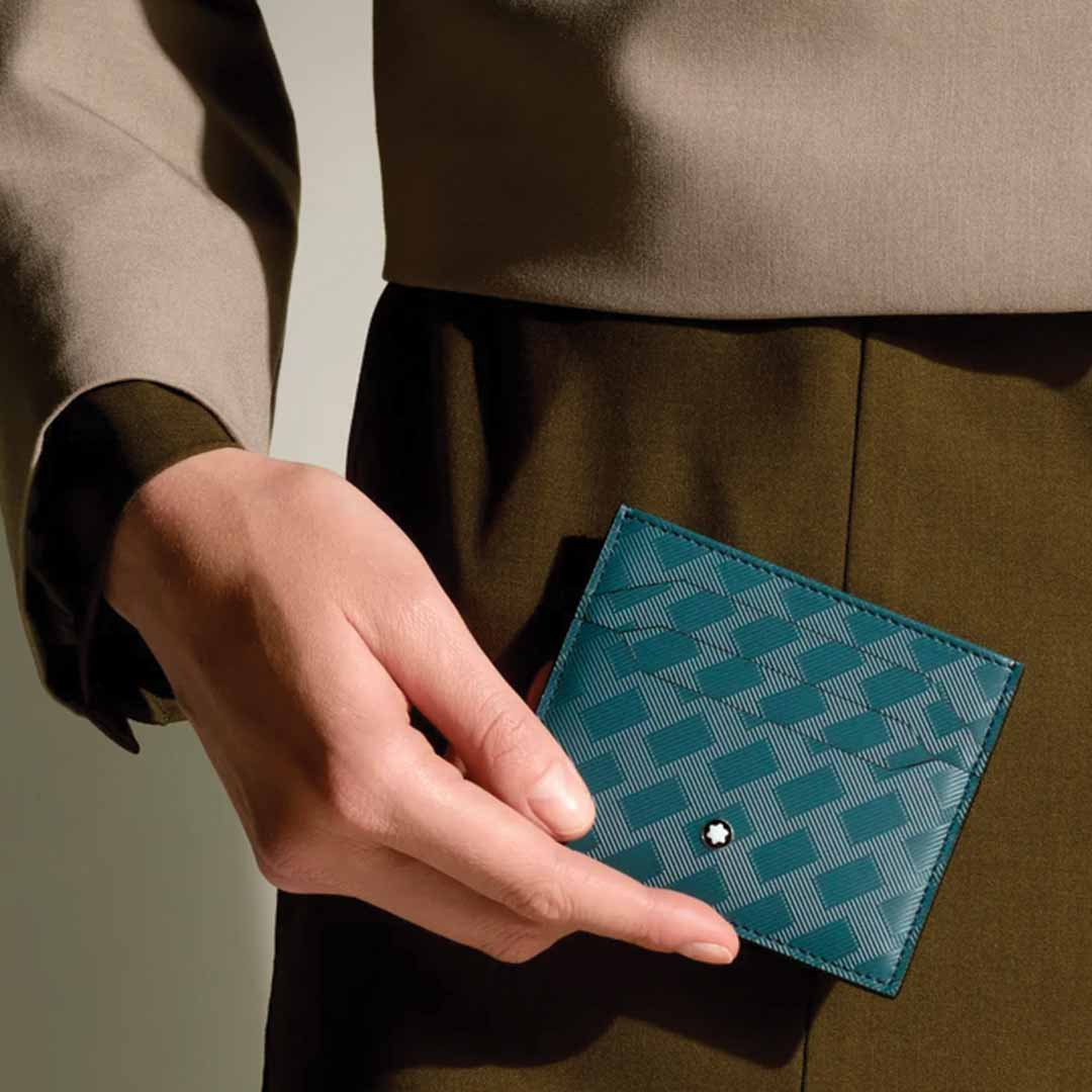 Dressed in a beige and olive ensemble, a person holds the Montblanc Extreme 3.0 6cc Leather Card Holder—a small, teal leather wallet featuring a textured pattern and adorned with a subtle round emblem in one corner, making it an elegant accessory for the stylish collection by Montblanc.