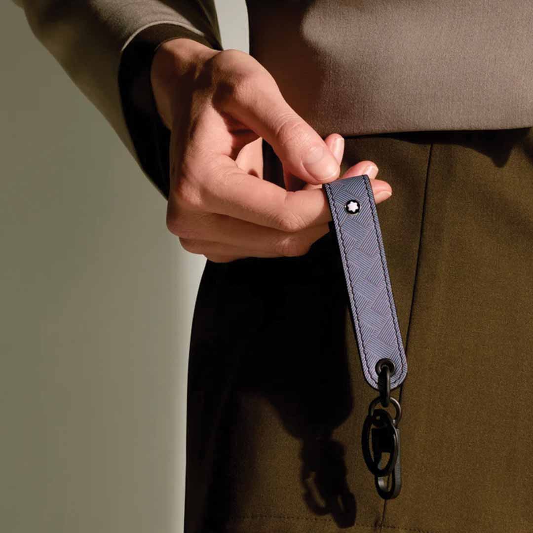 Dressed in a brown ensemble, an individual holds the Montblanc Extreme 3.0 Black Key FOB, which boasts a textured strap crafted from full-grain leather, a metal hook, and a small emblem. The softly lit background accentuates the contrast between the fabric and this elegant key fob from Montblanc.