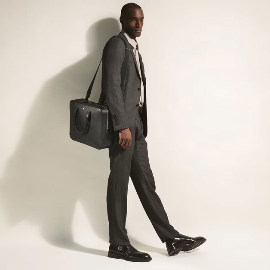 A man in a grey suit and black dress shoes walks carrying the Montblanc Sartorial Medium Leather Document Case Bag with a shoulder strap, set against a plain light background.