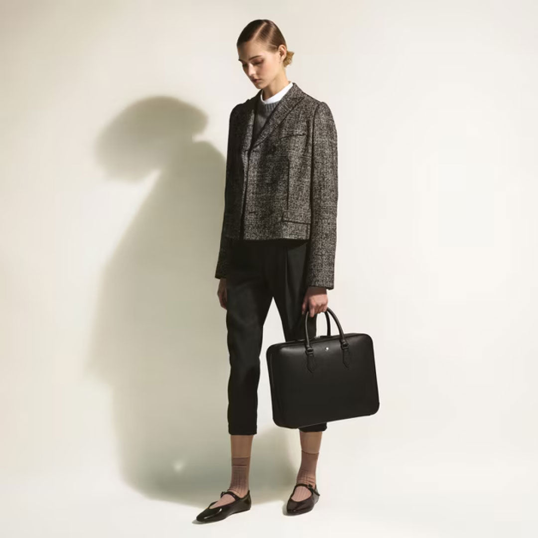 A person in a grey textured blazer, cropped black pants, and black Mary Jane shoes stands against a plain background, holding the Montblanc Sartorial Medium Leather Document Case Bag by Montblanc and looking downward.