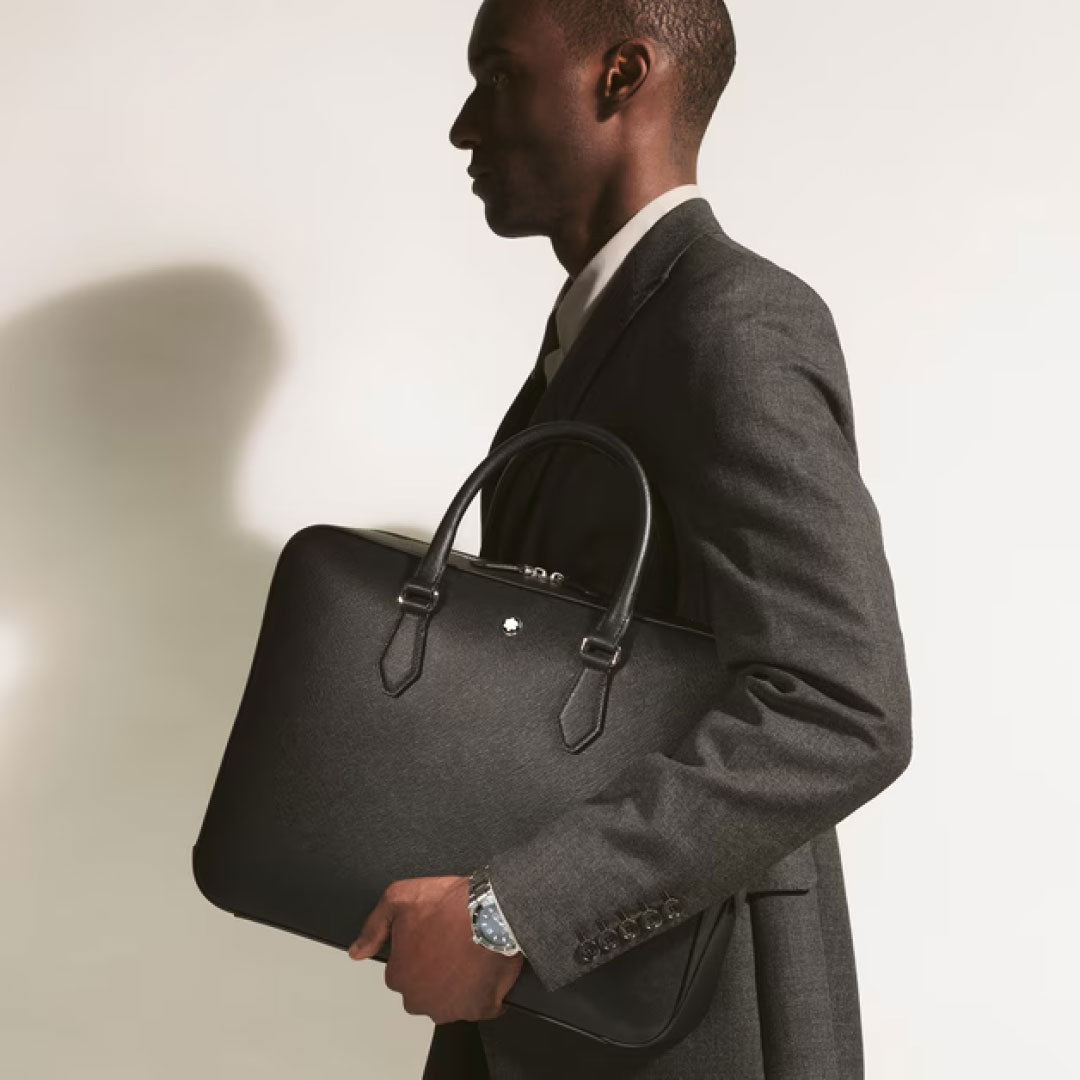 A man in a dark suit holds the Montblanc Sartorial Medium Leather Document Case Bag under his arm, standing against a plain, light-colored background.