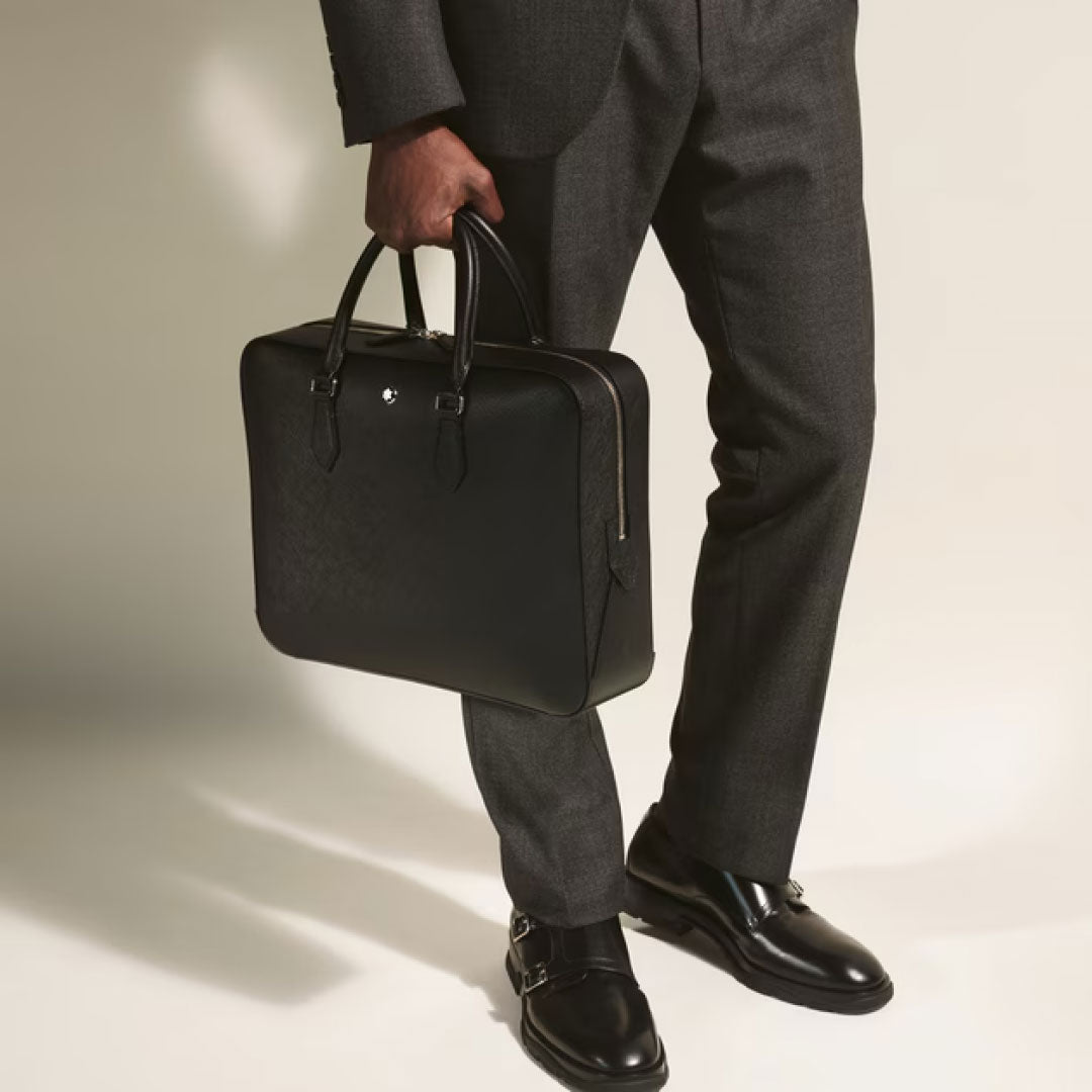A person in a dark gray suit holds the Montblanc Sartorial Medium Leather Document Case Bag by Montblanc and wears black double monk strap shoes, standing against a light background.