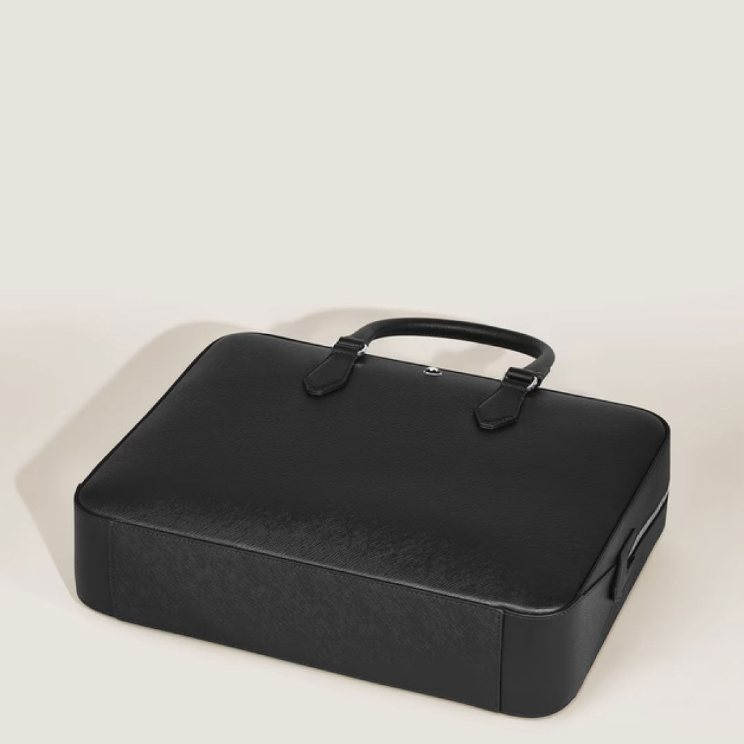 A Montblanc Sartorial Medium Leather Document Case Bag in black, featuring two handles, is shown closed on a light beige surface, photographed from an elevated angle.