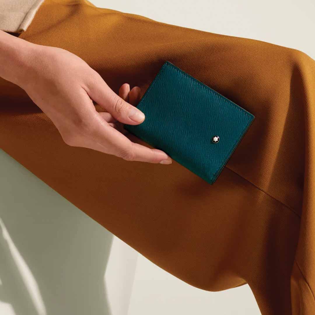 A person's hand holds the elegant Montblanc Sartorial 4cc Mini Leather Wallet in Cyprus blue, showcasing credit card slots and a subtle Montblanc logo, as it rests on their lap. The person is dressed in mustard-colored pants.
