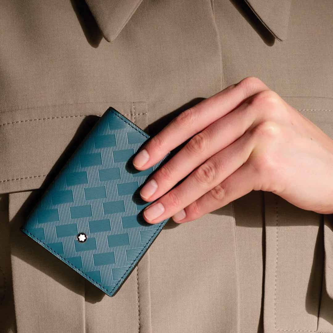A person holding a Montblanc Extreme 3.0 4cc Leather Card Holder in avio blue leather against a khaki-colored jacket. The wallet's subtle pattern and small circular emblem create a striking contrast with the clothing, as the hand is positioned near the chest to highlight this elegant accessory from Montblanc.