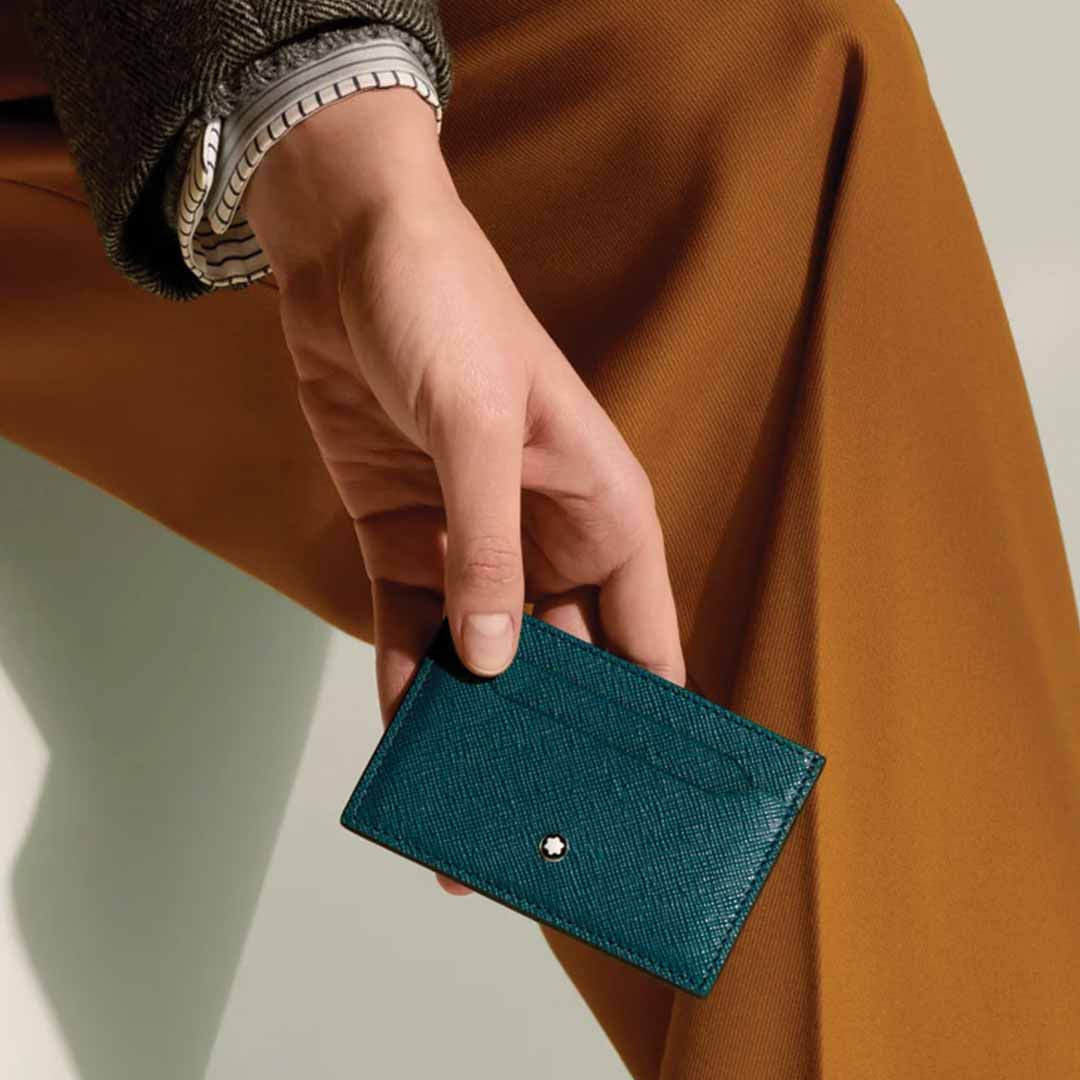 A person wearing a brown jacket and mustard pants holds a small, textured teal wallet made from Saffiano-printed leather. The Montblanc Sartorial 4cc Leather Card Holder is subtly adorned with the Montblanc emblem, set against a soft, neutral background.
