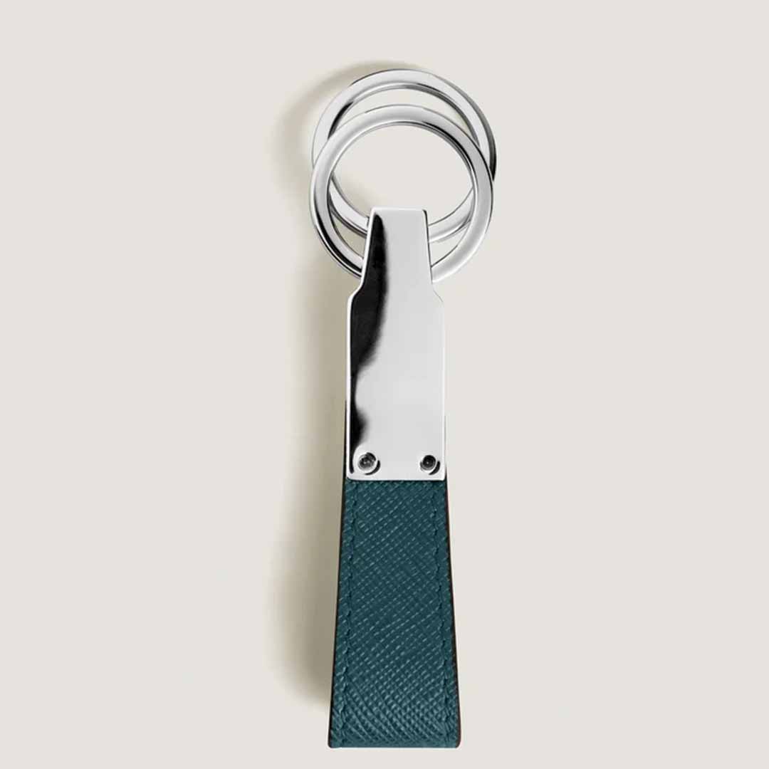 The Montblanc Sartorial Loop Key Fob showcases interlocking rings and a textured dark green leather strap. The rectangular strip, highlighted with Cyprus blue leather and secured by two small screws, hangs gracefully against a plain light background.