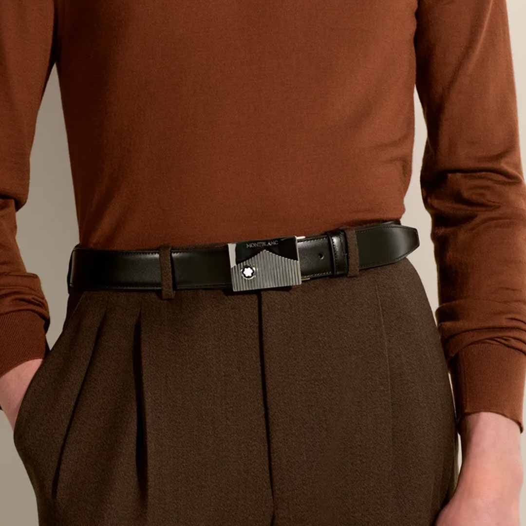 A person wearing a brown long-sleeve shirt tucked into brown pleated trousers is sporting a Montblanc Black 35mm Leather Belt with a rectangular, silver-accented buckle, featuring the iconic Montblanc logo in the center.