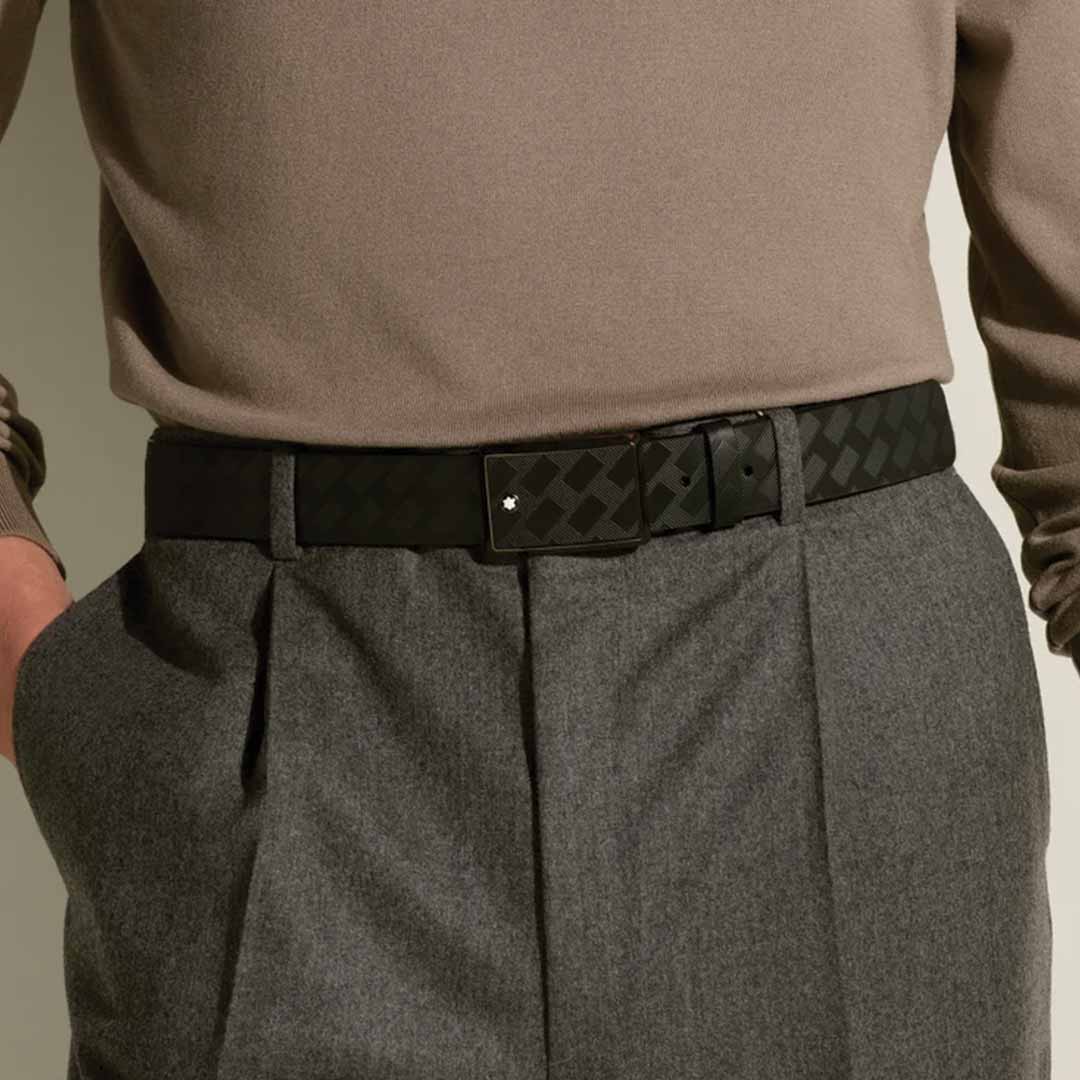 A person wearing a taupe long-sleeve top and gray pleated pants with hands in pockets, accessorized with the Montblanc 35mm Leather Belt featuring an Extreme 3.0 print.