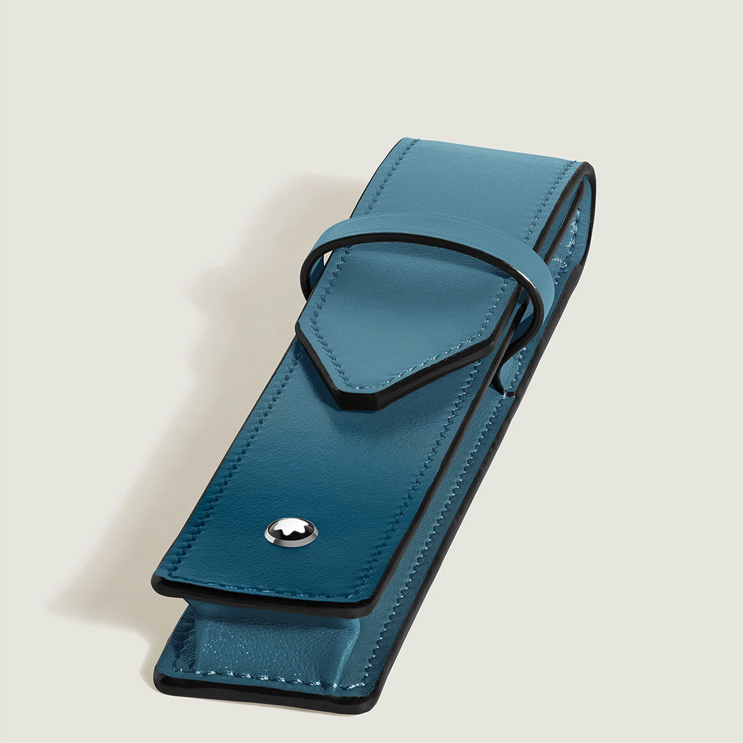 The Montblanc Meisterstück Leather Pen Pouch, crafted from blue full-grain calfskin with black edges and a flap closure, features a round silver Montblanc emblem on the front and is displayed against a plain light background.