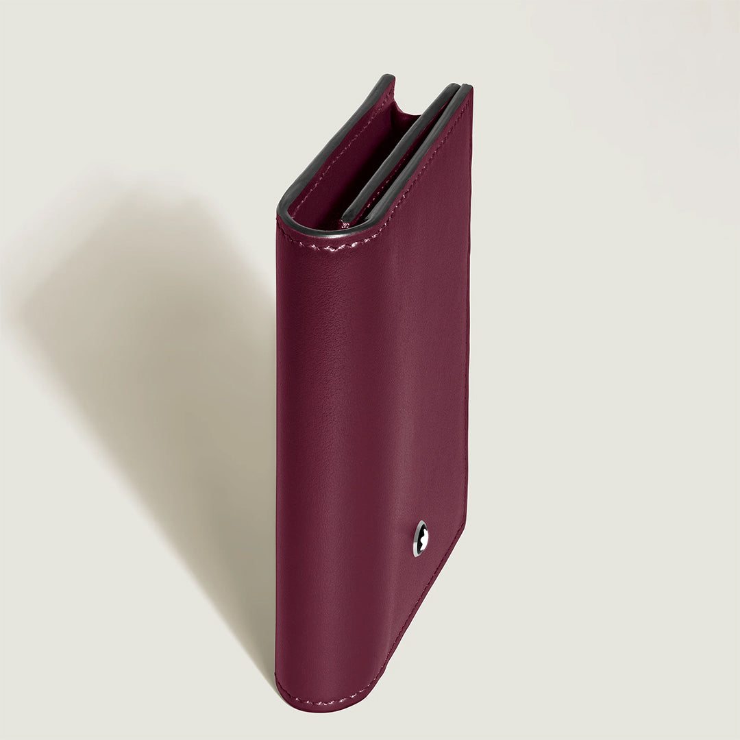 A vertical view of the Montblanc Meisterstuck 4CC Card Holder, closed in burgundy, stands upright on a light surface. Its minimalist design with a small round Montblanc emblem on the front is ideal for elegantly storing business cards.