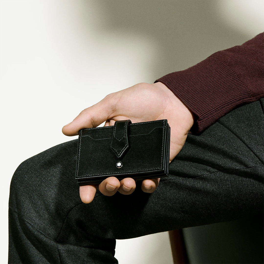 A person in a maroon sweater and dark pants holds the sleek Montblanc Meisterstück 5CC Card Holder. The cardholder's simple flap with button detail stands out against the neutral background, enhancing its minimalist Montblanc-inspired aesthetic.