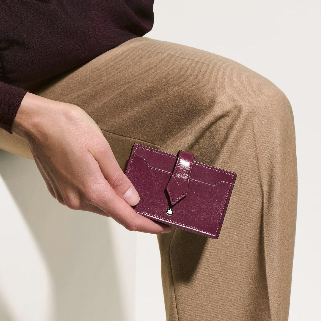 A person is holding a Montblanc Meisterstuck 5CC Card Holder. They're in tan pants and a dark long-sleeved shirt, seated on a light surface with a neutral background.