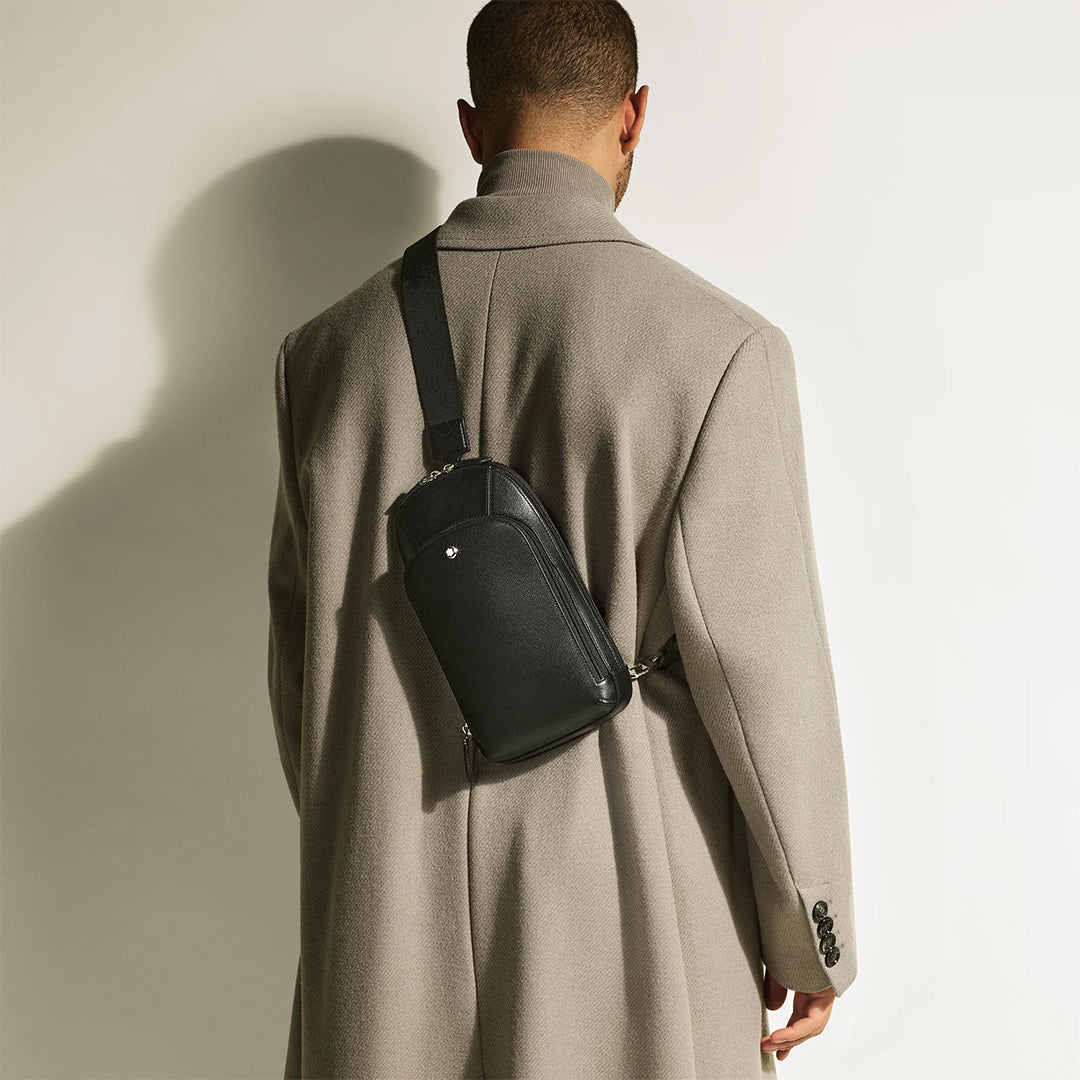 A person faces away, wearing a long beige coat and a Montblanc Sartorial Sling Bag slung across their back. The plain background softly shadows the figure, emphasizing the bag's adjustable shoulder strap.