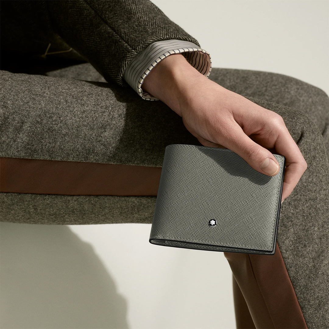 A person in a tailored grey and brown suit sits with one hand on their knee, holding the Montblanc Sartorial 8CC Leather Wallet, which features Saffiano-print leather, a white Montblanc emblem, and multiple credit card slots for practical elegance.