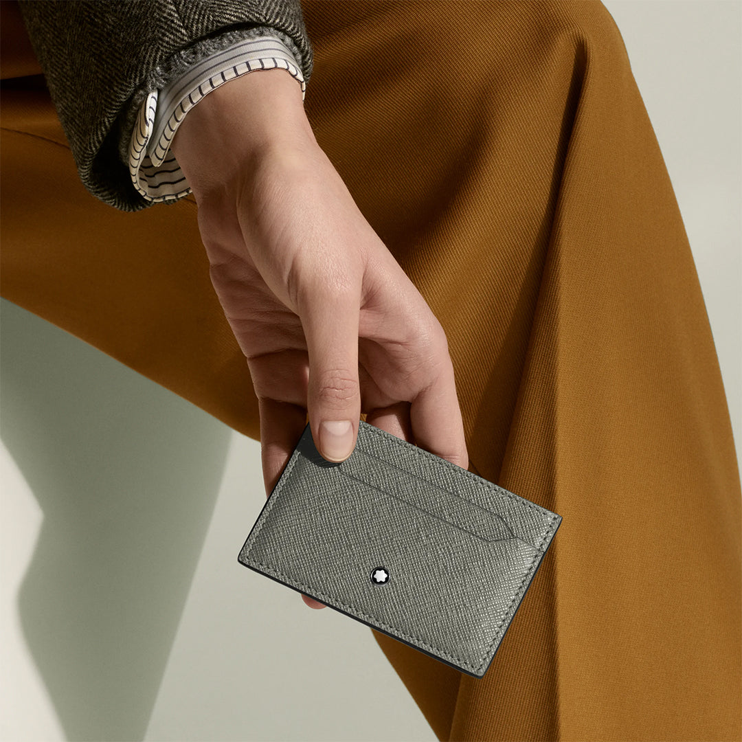A person in a brown suit and striped shirt holds the Montblanc Sartorial Grey 5CC Leather Card Holder, featuring a Saffiano print and the Montblanc emblem at the center.
