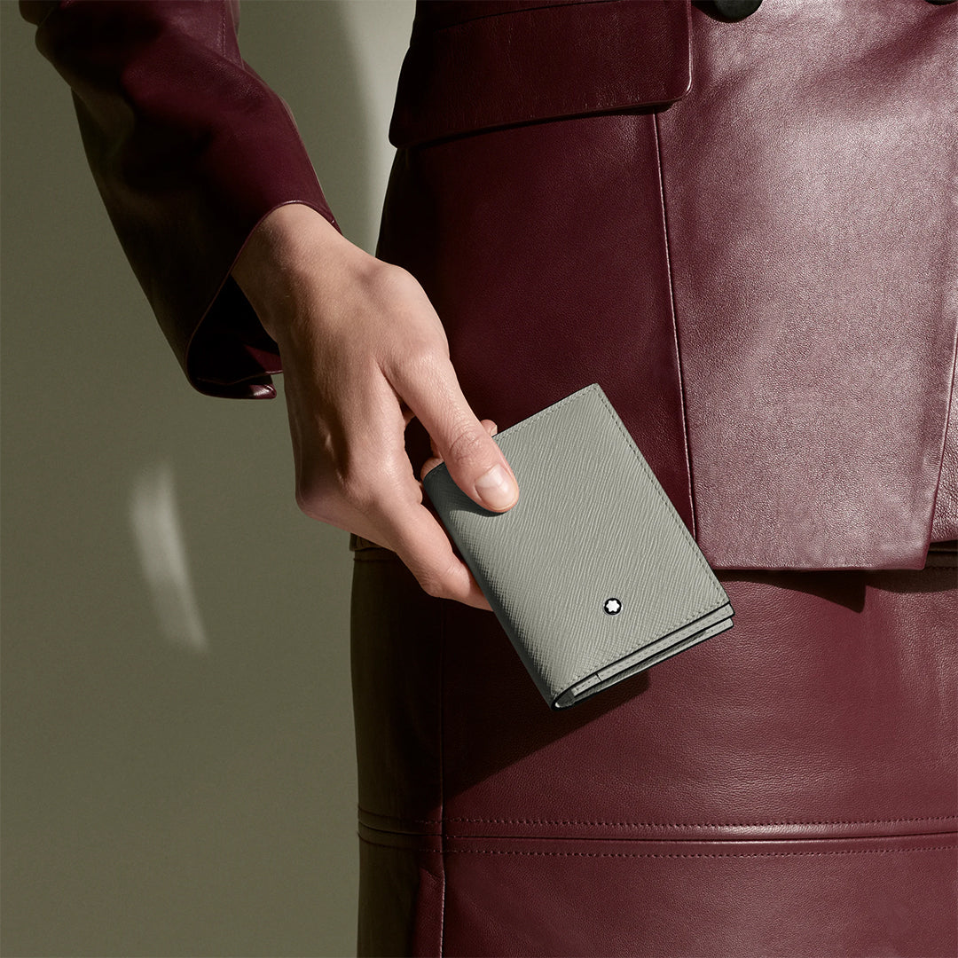 A person in a burgundy leather suit holds the Montblanc Sartorial 4CC Leather Card Holder, featuring gray Saffiano-printed leather and a round metallic Montblanc emblem, against a neutral background.