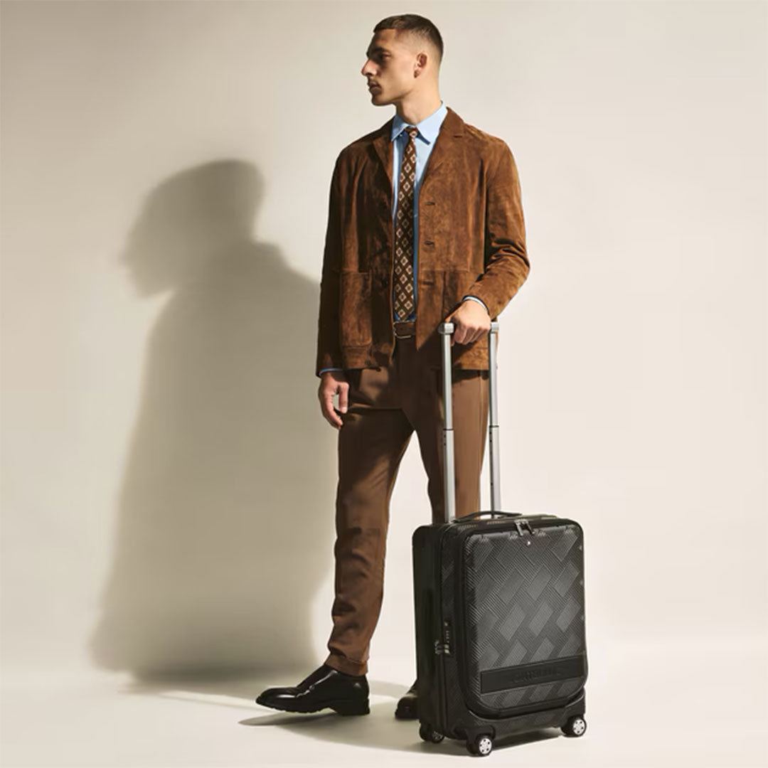 A man in a brown suit, blue shirt, and tie stands against a light background, holding the handle of a Montblanc Extreme 3.0 Cabin with Pocket trolley bag by his side.