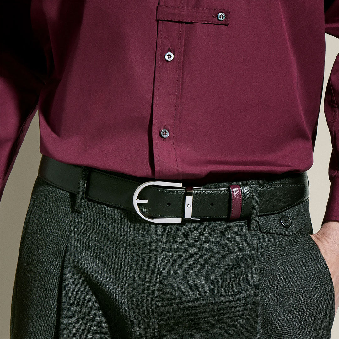 A person wearing a burgundy shirt and dark gray trousers pairs them with a Montblanc Horseshoe Buckle 35mm Reversible Leather Belt, featuring a silver buckle and burgundy accent. One hand is in their pocket.