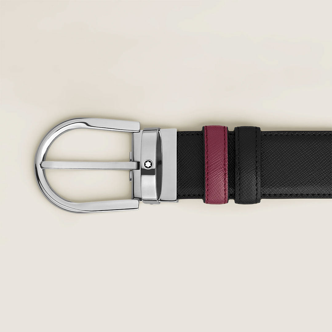 A close-up of the Montblanc Horseshoe Buckle 35mm Reversible Leather Belt, featuring black calf-skin leather with a shiny silver buckle and a dark red loop, displayed against a light beige background.