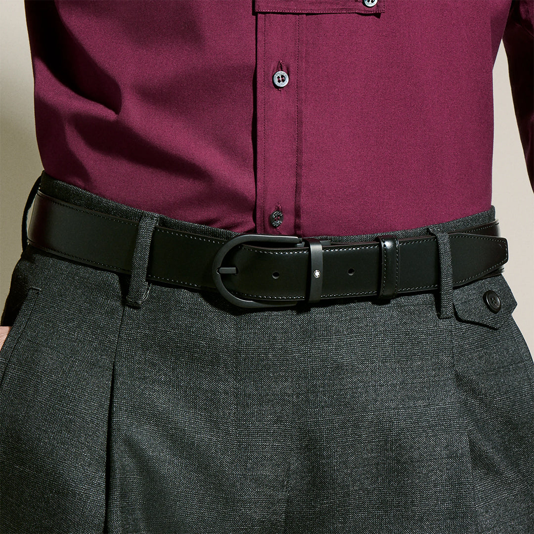 A person is wearing a maroon button-up shirt tucked into gray trousers, complemented by the Montblanc Horseshoe Buckle 35mm Black Leather Belt. Its simple horseshoe pin buckle adds subtle elegance, while the trousers have a side button detail.