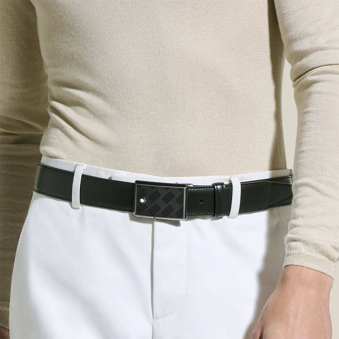 A person in a beige long-sleeve top and white pants highlights a Montblanc 35mm Black Leather Belt, featuring an Extreme 3.0 motif on its patterned buckle. The plain backdrop beautifully emphasizes the belt and ensemble.