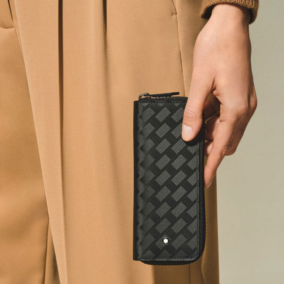 A person in tan pants and a brown sweater holds the Montblanc Extreme 3.0 Black Leather Double Pen Pouch, featuring a geometric pattern and a small white Montblanc emblem near the bottom.