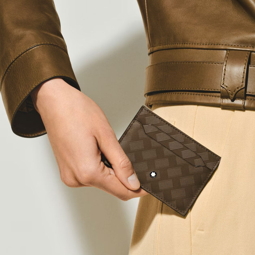A person in a brown leather jacket and beige pants holds a Montblanc Extreme 3.0 5cc Leather Card Holder featuring the brand’s signature small star emblem near the corner.