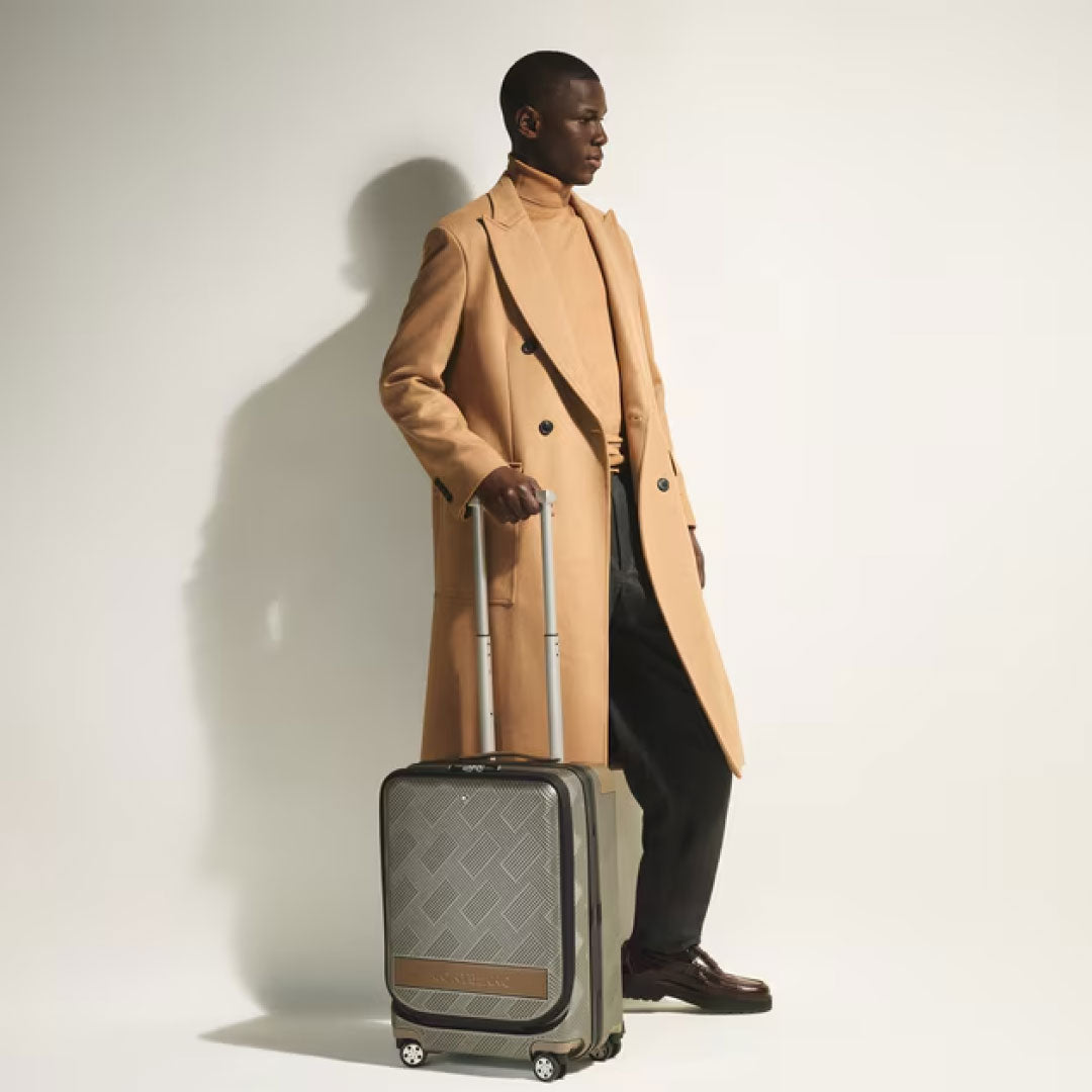 A man in a tan overcoat and turtleneck stands sideways in a minimalist setting, holding the handle of a Montblanc Extreme 3.0 Cabin Luggage with Pocket, his dark pants and shoes contrasting against the light background.