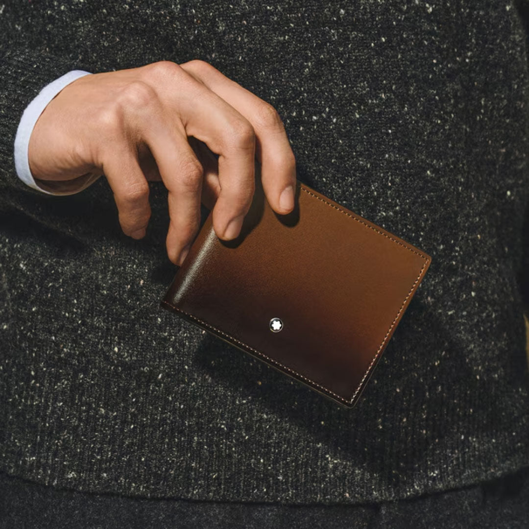 A person in a dark, speckled sweater holds the Montblanc Meisterstück Sfumato 6CC Leather Wallet, featuring the iconic metallic Montblanc emblem—a refined luxury accessory.