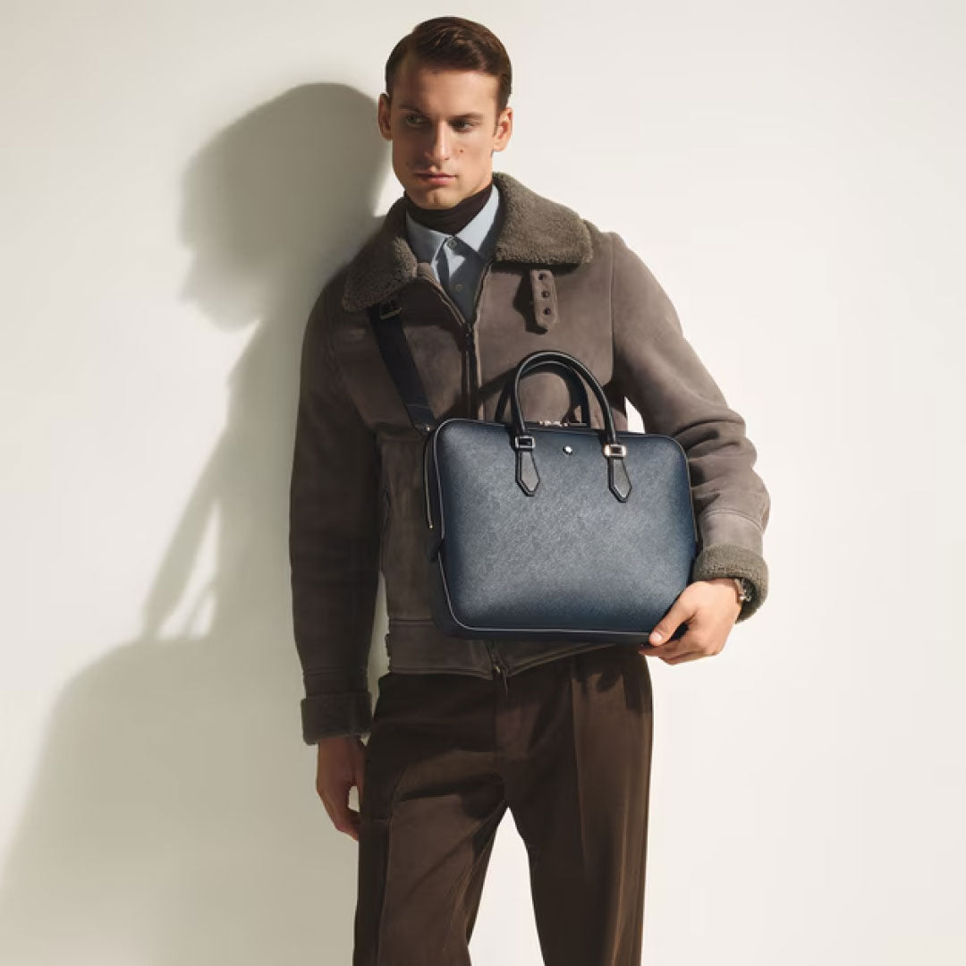 A man in a brown shearling-collared jacket and pants stands against a white background, holding the Montblanc Sartorial Thin Document Case Bag by Montblanc—a navy-blue business briefcase with a shoulder strap.