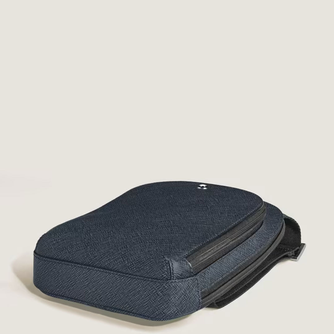 A dark blue Montblanc Sartorial Leather Sling Bag with a textured finish, zipper, and refined leather details lies flat on a light beige surface. A small white Montblanc logo appears near the top.