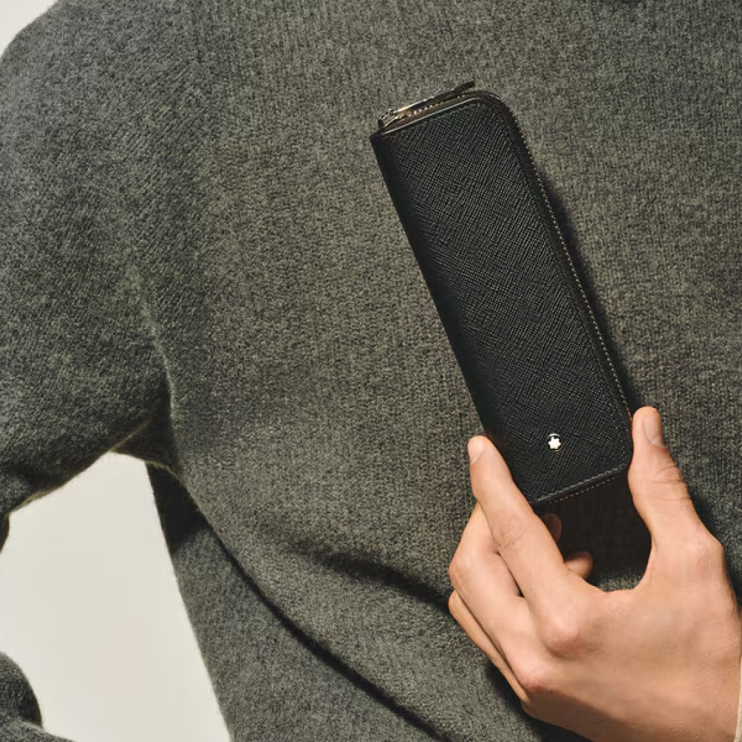 A person in a gray sweater holds a Montblanc Sartorial Leather 2 Pen Pouch With Zip, featuring the brand's white star logo, upright against their chest.