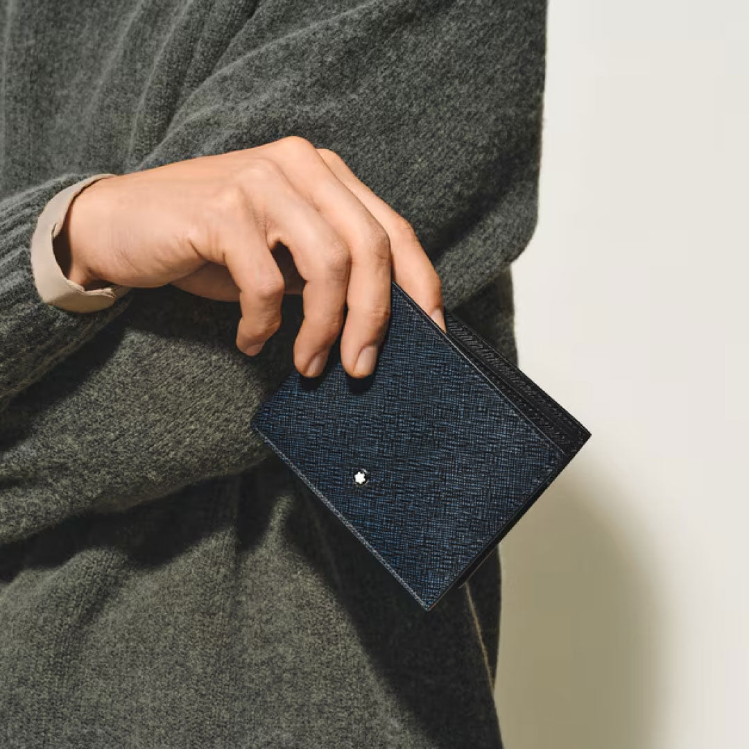 A person in a grey sweater holds the Montblanc Sartorial Leather 6cc Wallet, featuring a small metallic Montblanc emblem at the center, set against a plain light background.