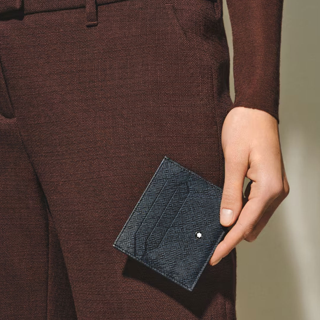 A person in a brown textured outfit holds the Montblanc Sartorial Leather 6cc Card Holder, featuring a small metallic emblem, with focus on the hand and the elegant Montblanc card holder set against a neutral background.