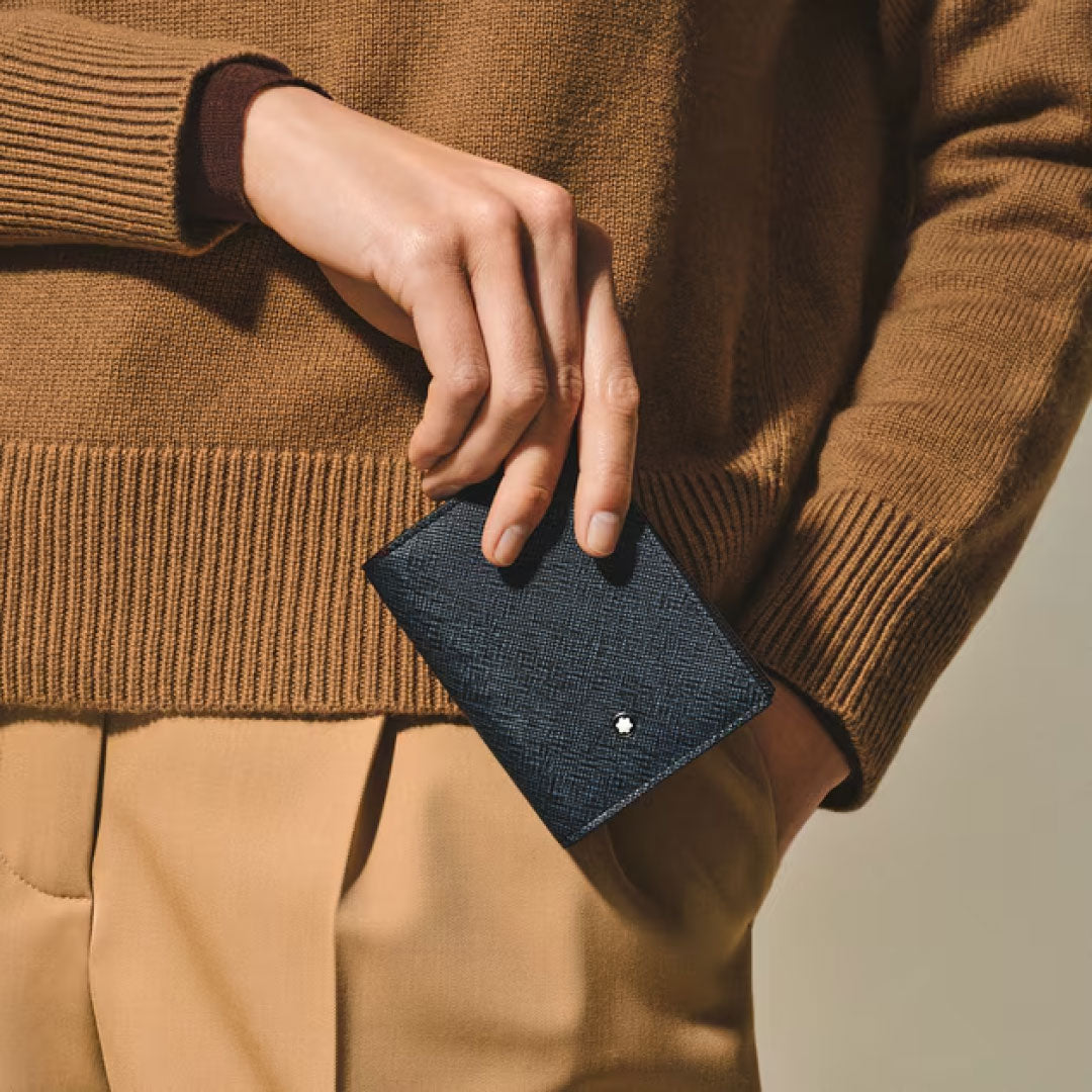 A person in a brown sweater and tan pants holds the Montblanc Sartorial Leather 4cc Card Holder, displaying its organized credit card slots and the iconic Montblanc white emblem near a pocket.
