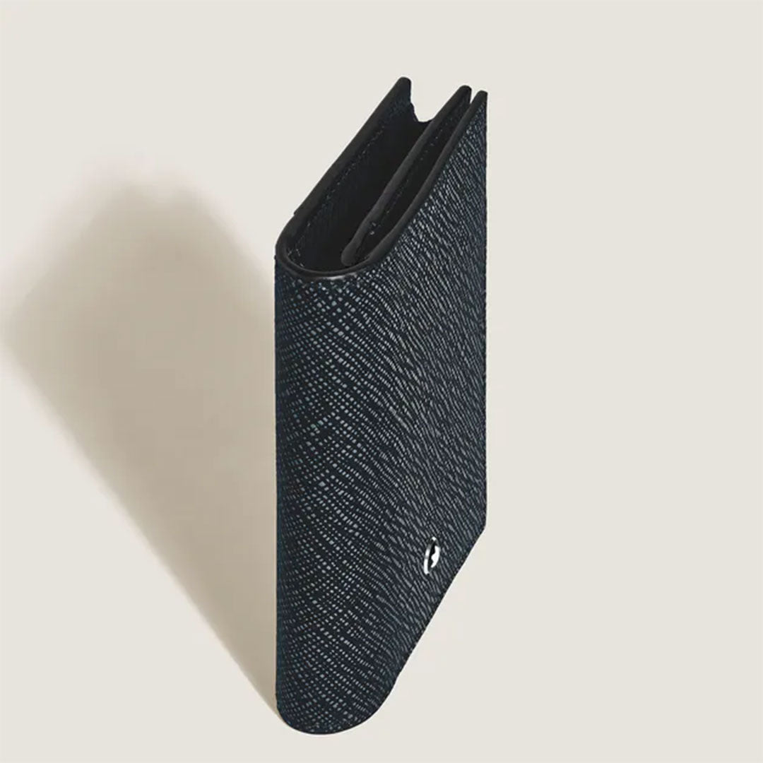 The Montblanc Sartorial Leather 4cc Card Holder stands upright on a light beige surface, casting a left shadow. This black Montblanc wallet is closed, with a metallic logo at the bottom corner and features multiple interior credit card slots.