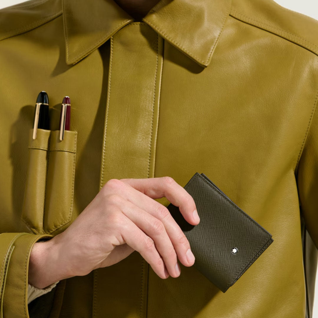 A person in a green Montblanc Sartorial leather jacket with pen holders holds a small, dark green Montblanc Sartorial 4CC Leather Card Holder featuring credit card slots and a round silver emblem.