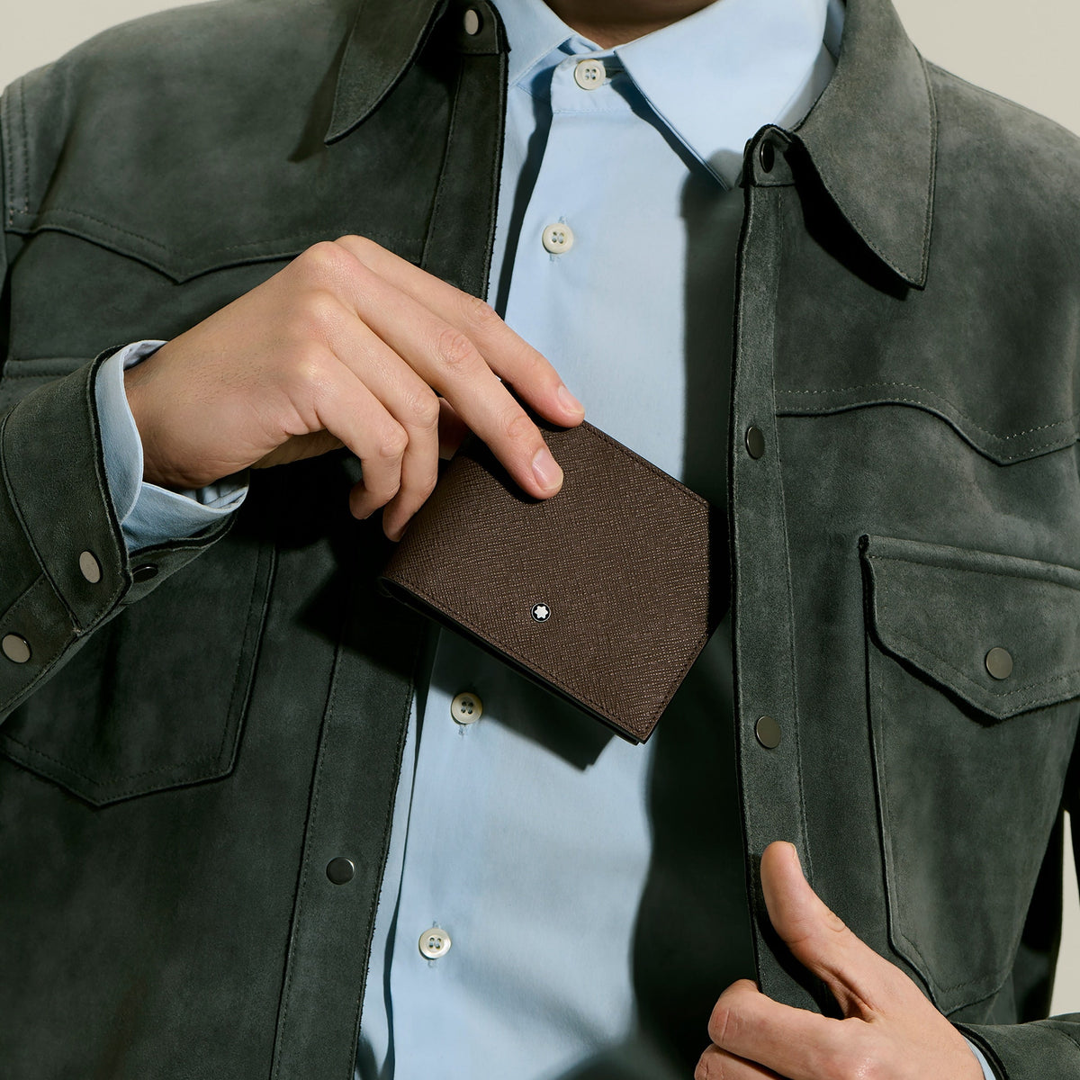 A person in a light blue shirt and dark jacket holds a Montblanc Sartorial 6CC Leather Wallet by Montblanc, featuring an embossed leather finish and small white logo, partially tucked into their jacket.